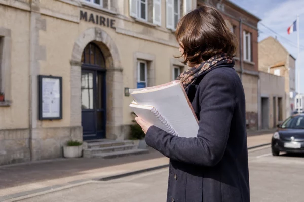 Un collaborateur municipal anonyme tient un dossier devant une mairie française, avec une rue calme en arrière-plan.