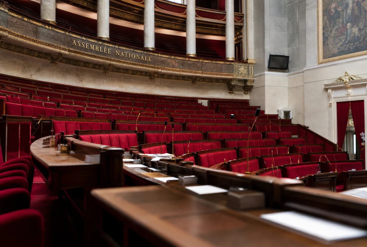 Intérieur de l’Assemblée nationale, sièges rouges vides et micros de séance dans une lumière naturelle claire.