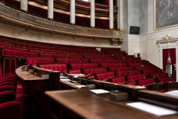 Intérieur de l’Assemblée nationale, sièges rouges vides et micros de séance dans une lumière naturelle claire.