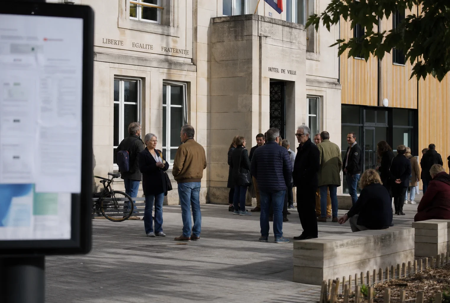 Scène de vie citoyenne à Aubervilliers devant une mairie, avec passants anonymes et ambiance politique locale.