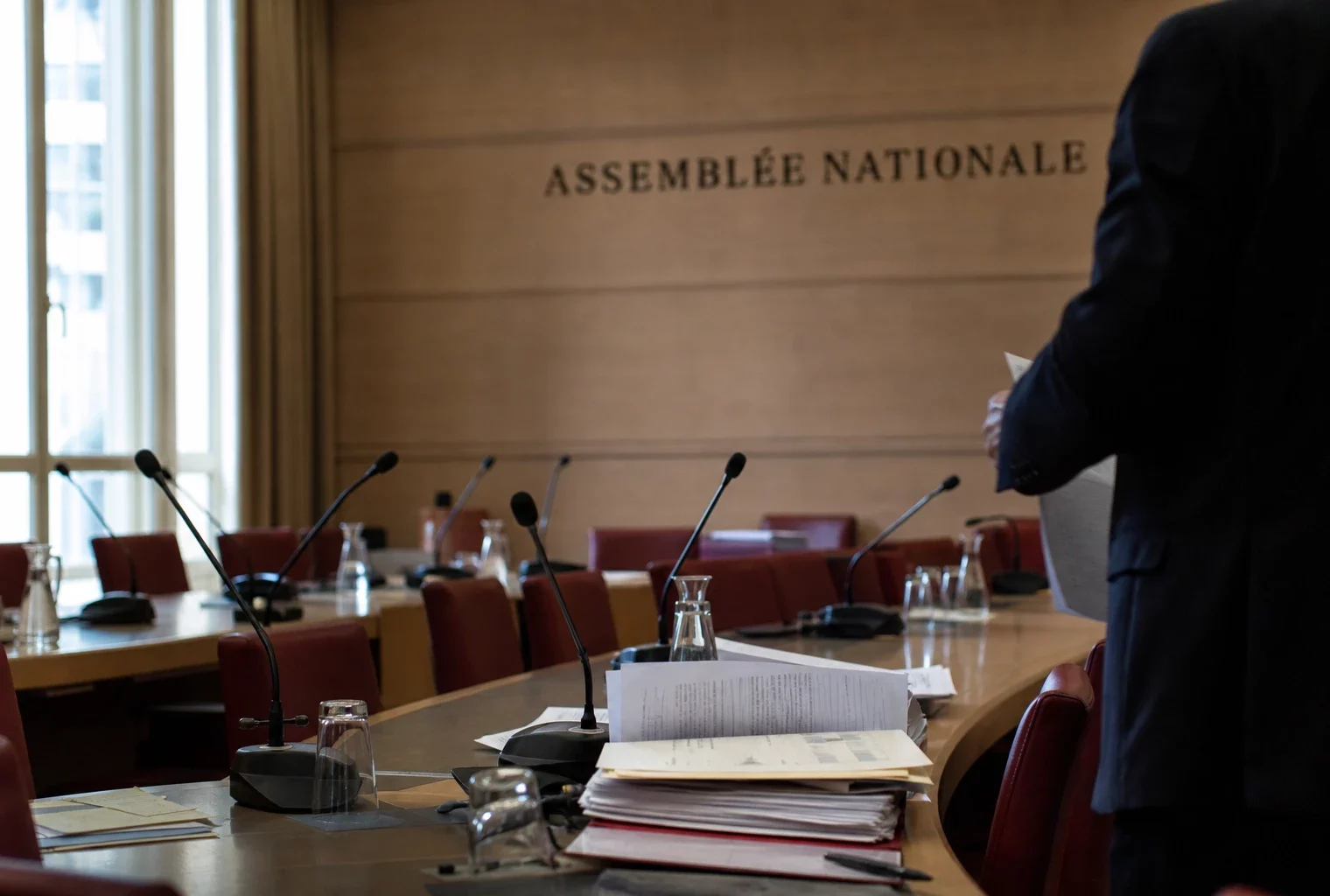 Salle d’audition à l’Assemblée nationale avec micros, dossiers ouverts et silhouettes anonymes en lumière naturelle.