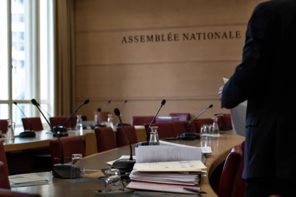 Salle d’audition à l’Assemblée nationale avec micros, dossiers ouverts et silhouettes anonymes en lumière naturelle.
