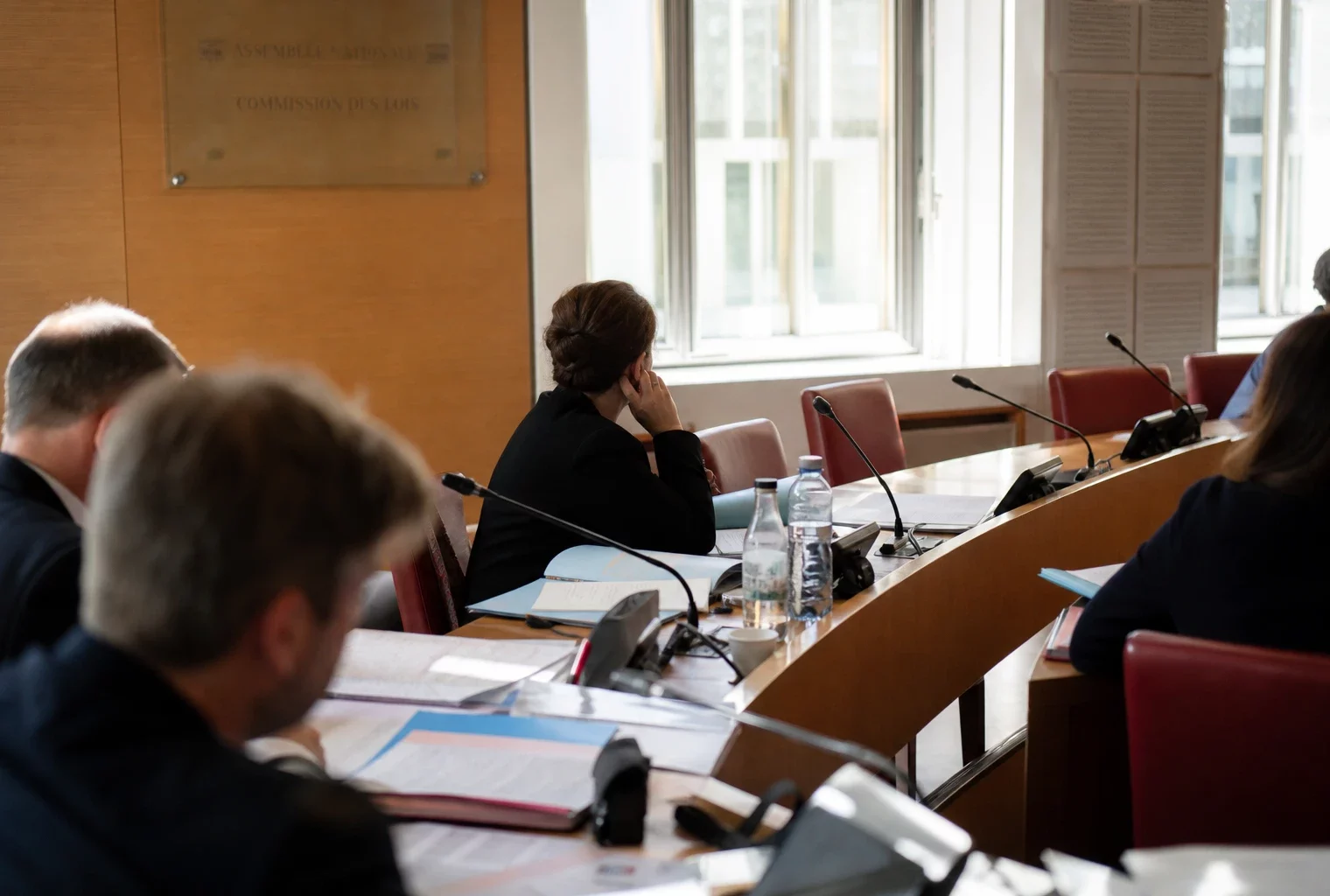 Audition en commission à l’Assemblée nationale avec micros, dossiers ouverts et silhouettes anonymes sous une lumière claire.
