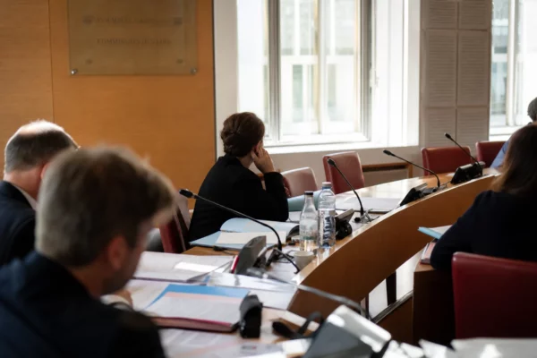 Audition en commission à l’Assemblée nationale avec micros, dossiers ouverts et silhouettes anonymes sous une lumière claire.