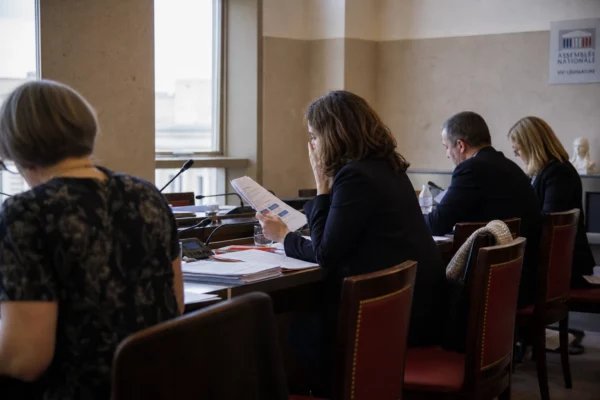 Audition parlementaire à l’Assemblée nationale avec micros, dossiers ouverts et silhouettes anonymes autour d’une table.