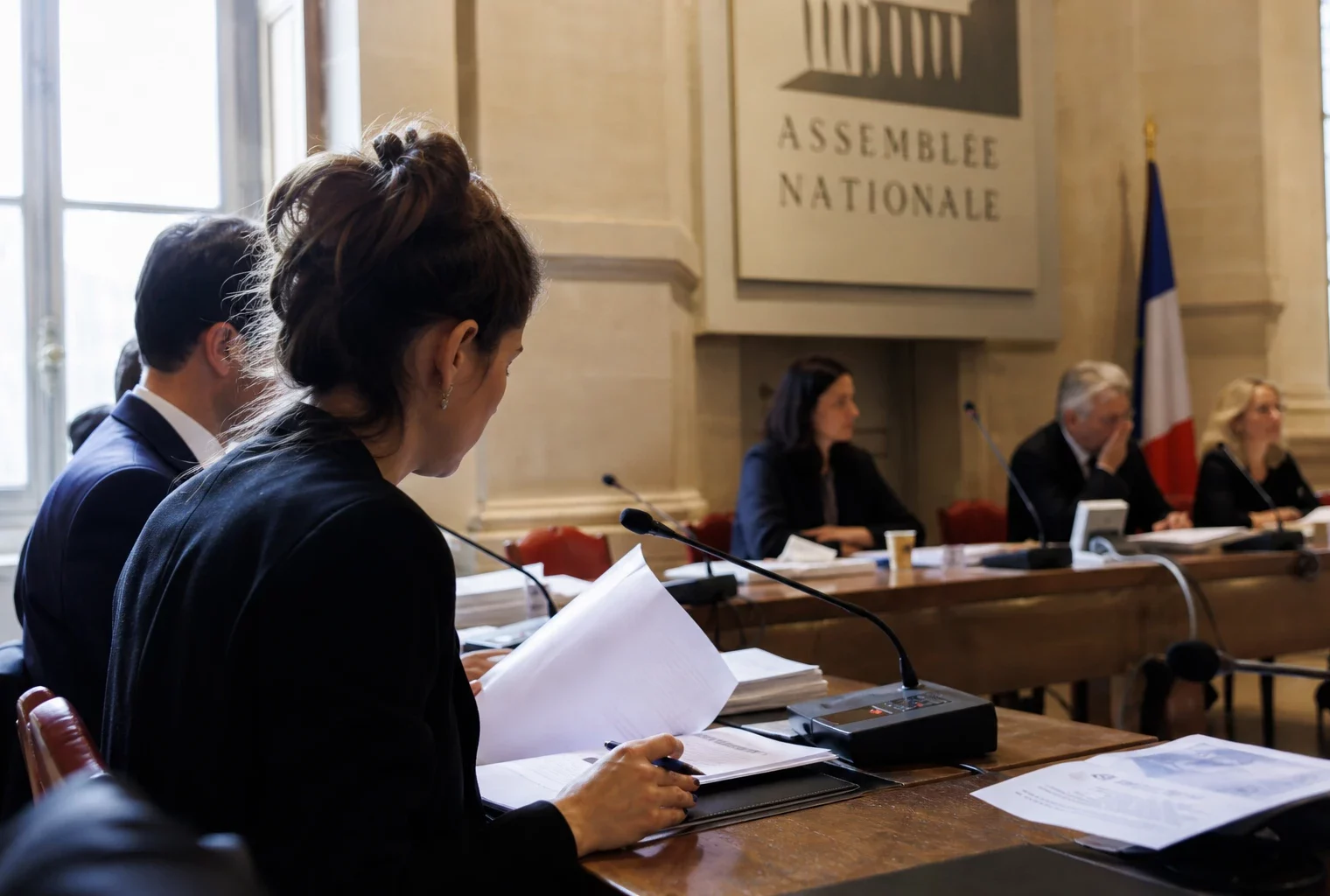 Salle d’audition institutionnelle lumineuse avec micros, dossiers ouverts et silhouettes anonymes en comité.