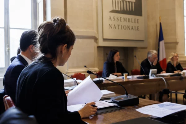 Salle d’audition institutionnelle lumineuse avec micros, dossiers ouverts et silhouettes anonymes en comité.