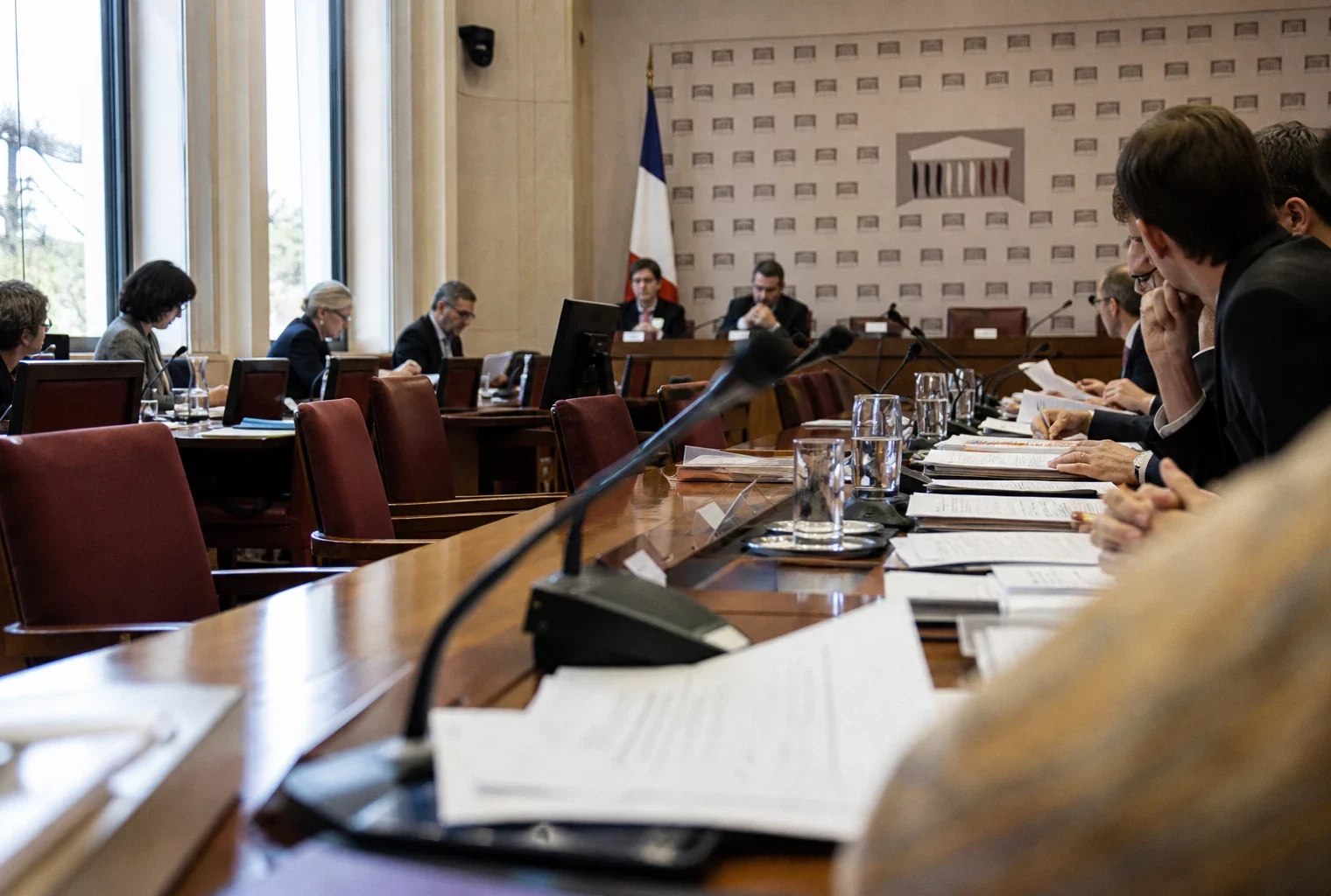 Audition parlementaire en salle claire avec micros, dossiers ouverts et silhouettes anonymes dans un décor institutionnel français.