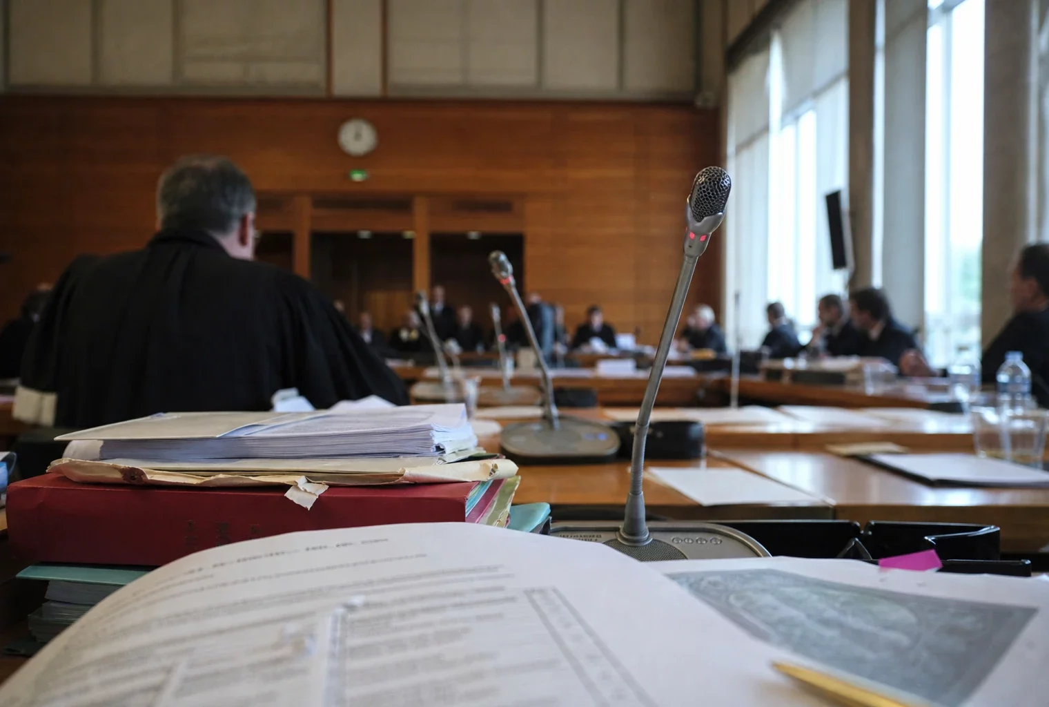 Audition institutionnelle en France avec micros de table, dossiers ouverts et silhouettes anonymes sous une lumière claire.
