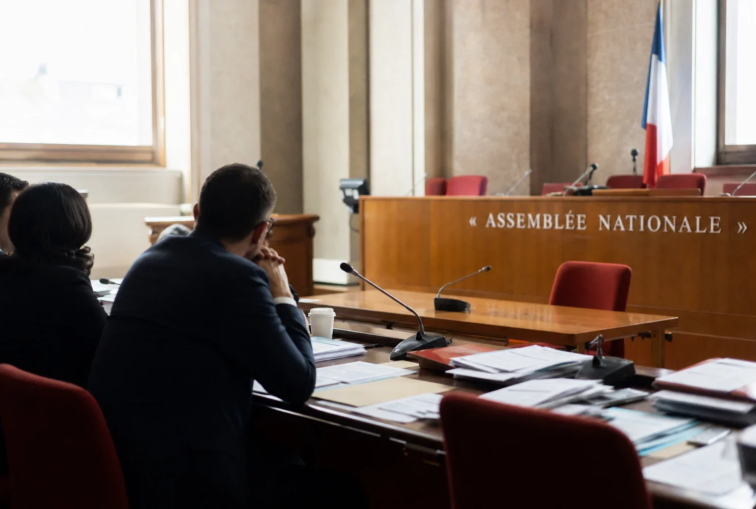 Salle d’audition parlementaire à l’Assemblée nationale avec micros, dossiers et silhouettes anonymes dans une lumière claire.