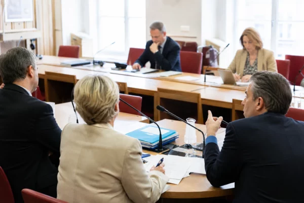 Réunion de commission parlementaire avec micros, dossiers ouverts et silhouettes anonymes sous une lumière claire.