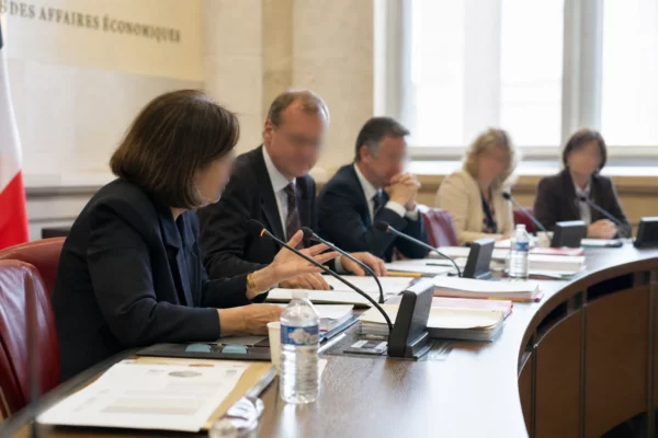 Audition parlementaire en commission avec micros, dossiers et silhouettes anonymes dans une salle française lumineuse.