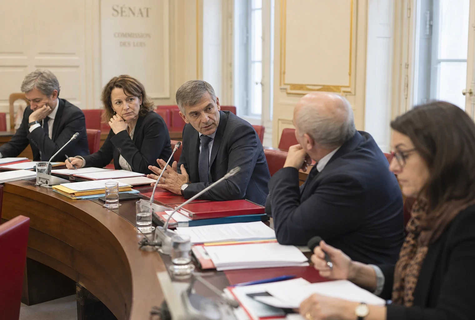 Réunion de commission au Sénat avec plusieurs responsables anonymes, micros, dossiers ouverts et lumière naturelle.
