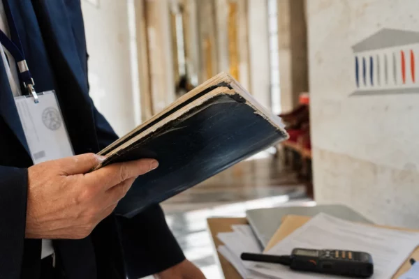 Dossier parlementaire tenu en main avec badge institutionnel et radio, dans un couloir lumineux de l’Assemblée nationale.