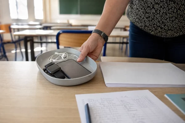 Main d’un surveillant posant un smartphone et des oreillettes confisqués sur une table d’examen au lycée