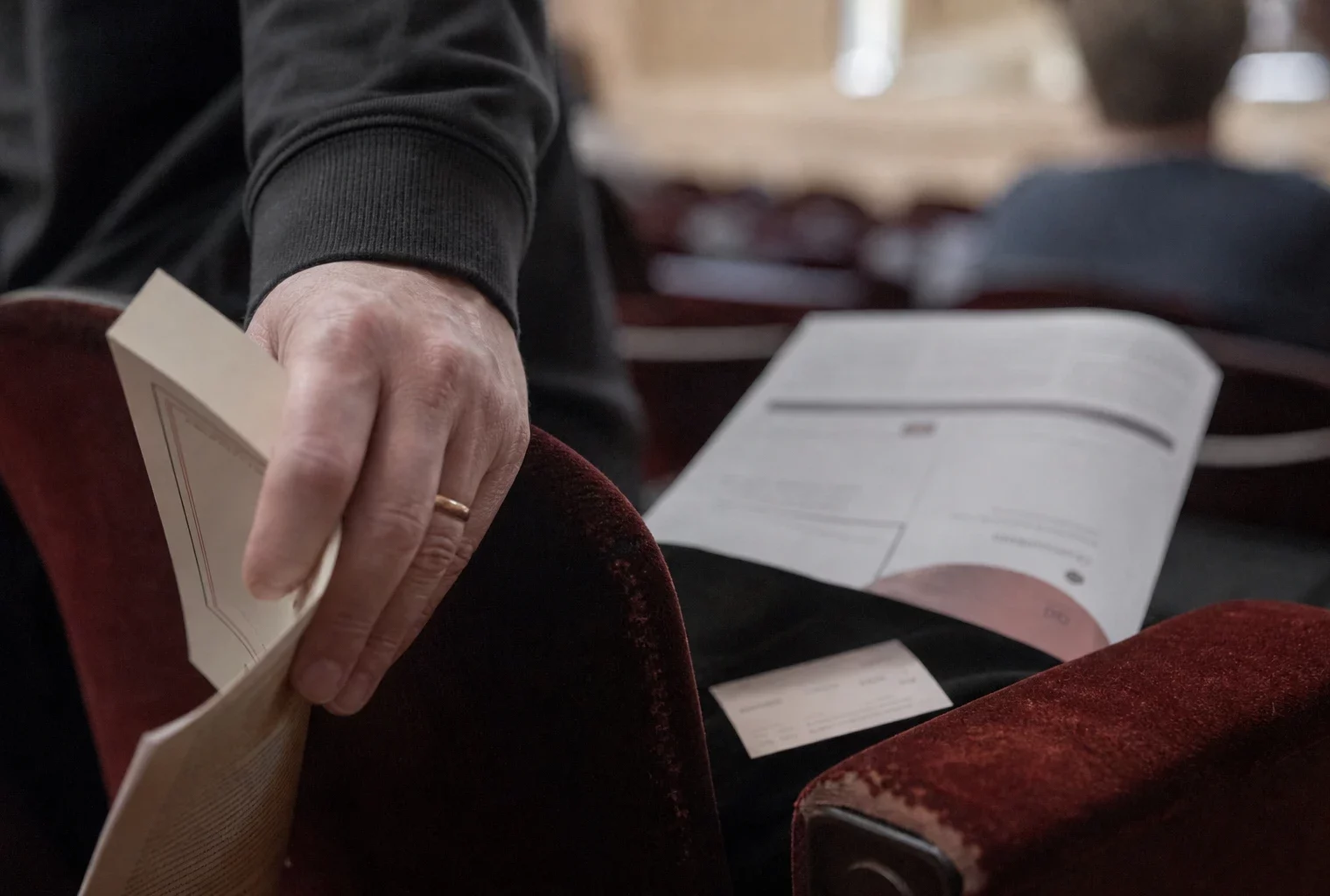 Gros plan d’une main sur un fauteuil de salle, avec un programme culturel flou et un livre avant une rencontre publique.
