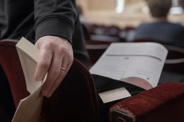 Gros plan d’une main sur un fauteuil de salle, avec un programme culturel flou et un livre avant une rencontre publique.