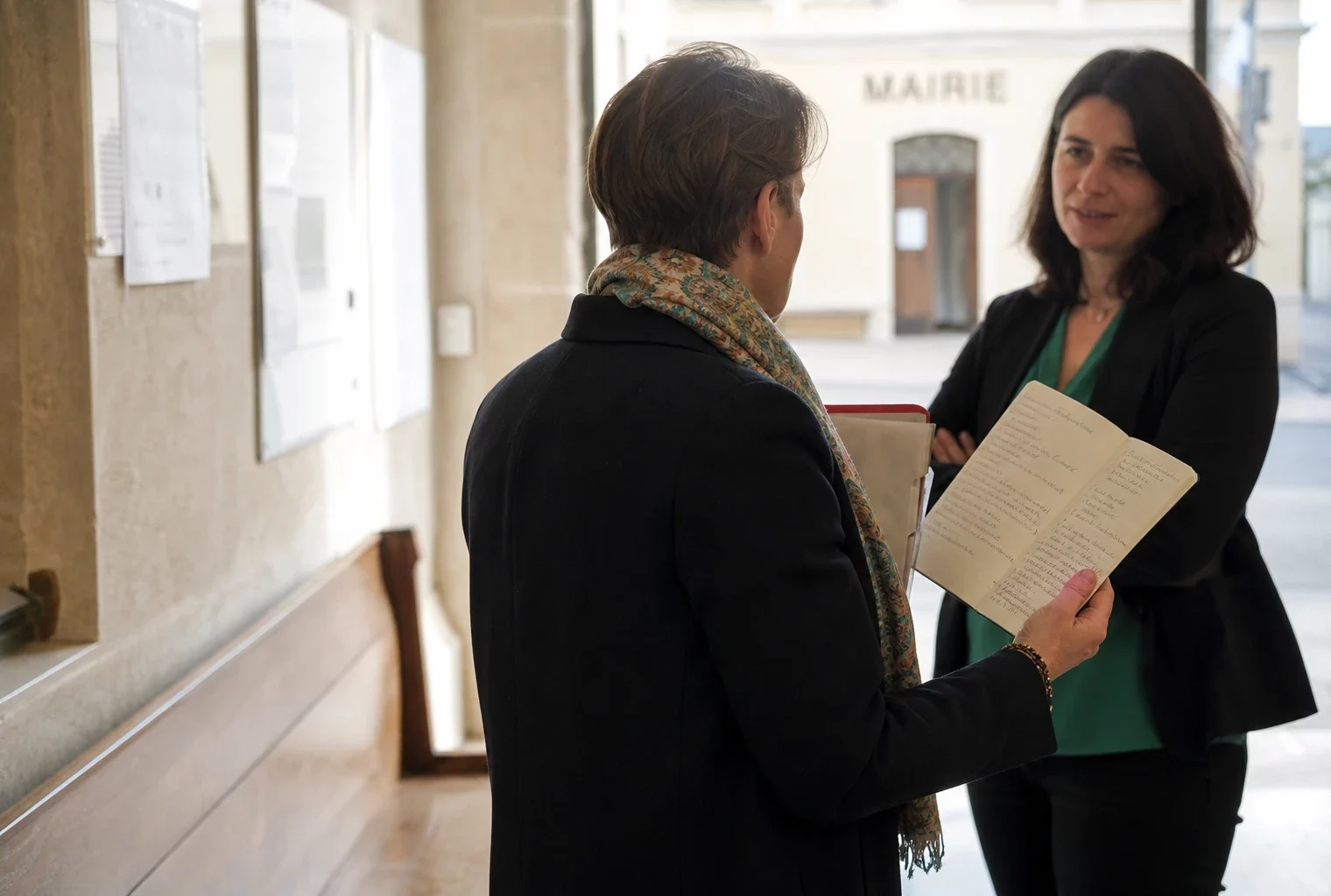 Un élu local anonyme échange dans un couloir de mairie avec un carnet et un dossier à la main.