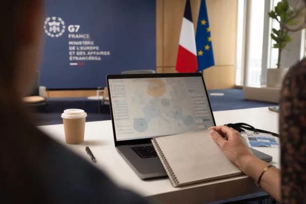 Vue de dos sur un bureau de briefing avec un écran affichant une interface européenne abstraite, en lumière naturelle claire.