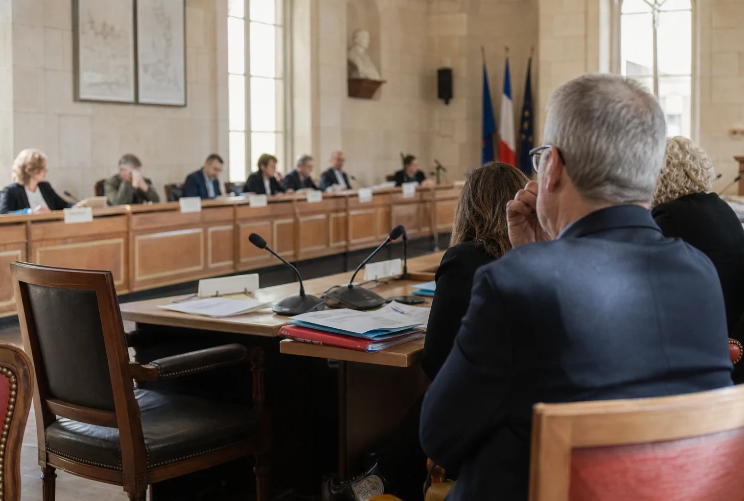 Salle municipale en plan large avec chaise vide, micros et dossiers flous, ambiance de budget local sous tension.