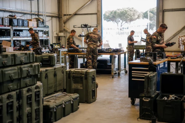 Techniciens et soldats dans un atelier militaire français inspectent du matériel et des caisses d’équipement sous une lumière claire.
