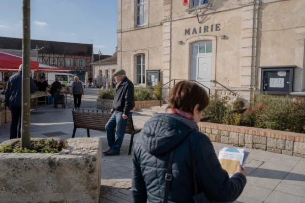 Devant une mairie de ville moyenne, des habitants anonymes traversent une place en plein jour.