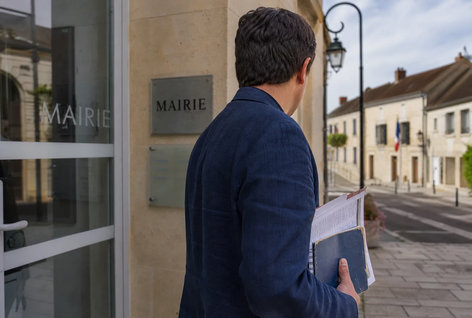 Un collaborateur local de dos, carnet à la main, devant une mairie française par forte chaleur.