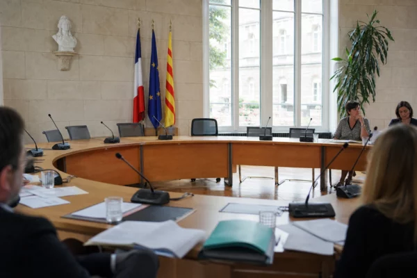 Salle municipale lumineuse avec micros, dossiers flous et chaise vide lors d’un briefing sur la canicule.