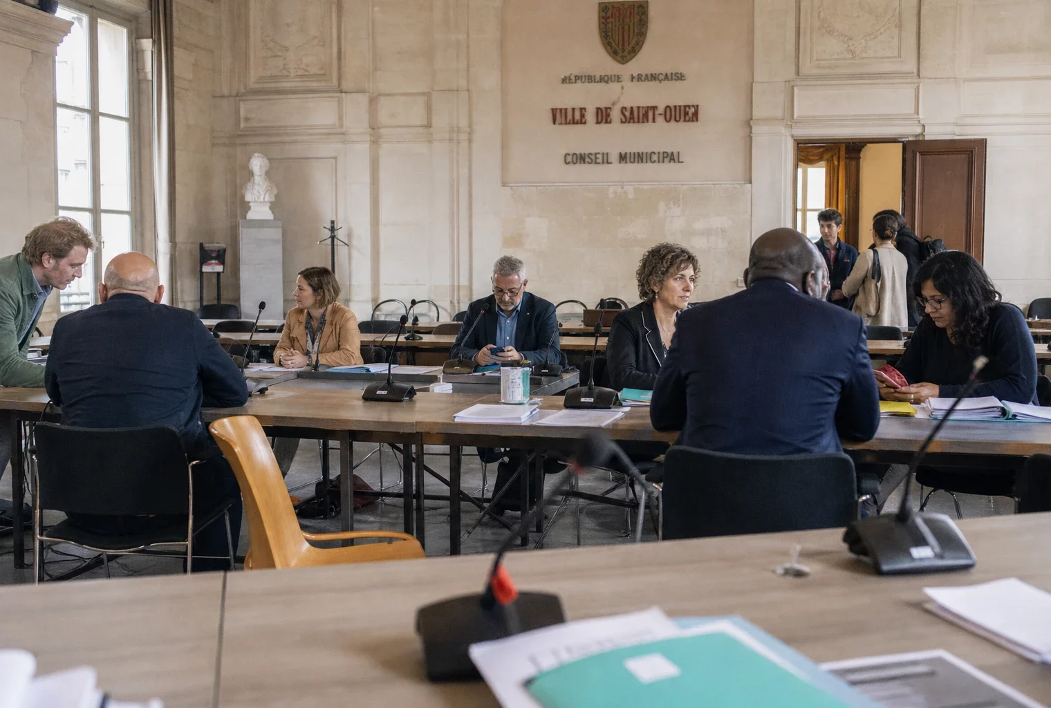 Salle municipale française avec micros, dossiers et chaise vide pendant un débat sur la canicule et le logement.