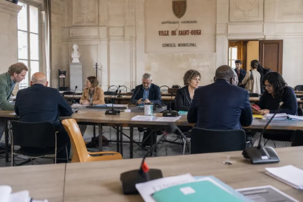 Salle municipale française avec micros, dossiers et chaise vide pendant un débat sur la canicule et le logement.