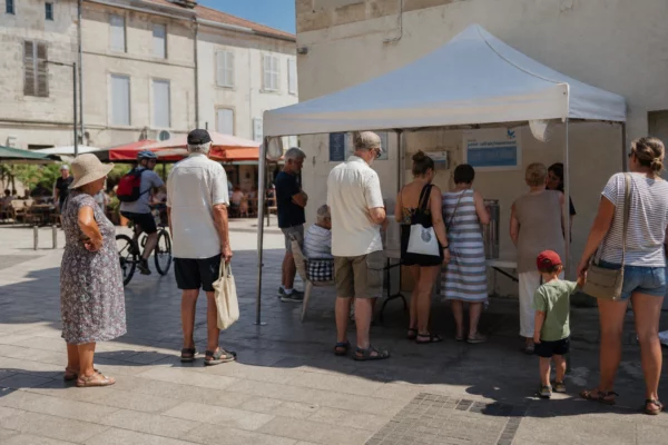 Place de ville française animée en pleine canicule, avec habitants à l’ombre et point d’eau municipal.