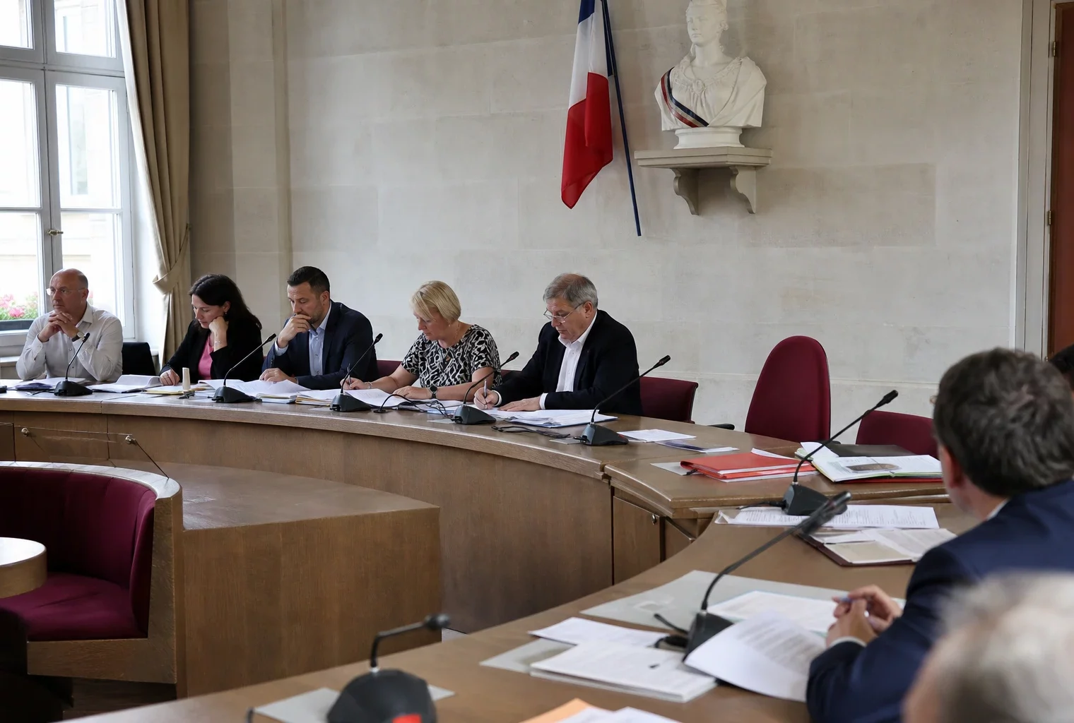 Salle municipale lumineuse avec micros, dossiers et une chaise vide lors d’une réunion politique locale.