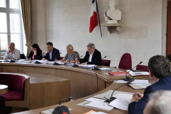 Salle municipale lumineuse avec micros, dossiers et une chaise vide lors d’une réunion politique locale.