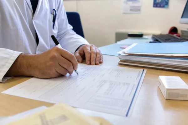 Main écrivant sur un dossier médical et judiciaire flouté dans un bureau de consultation français, avec médicaments anonymes.