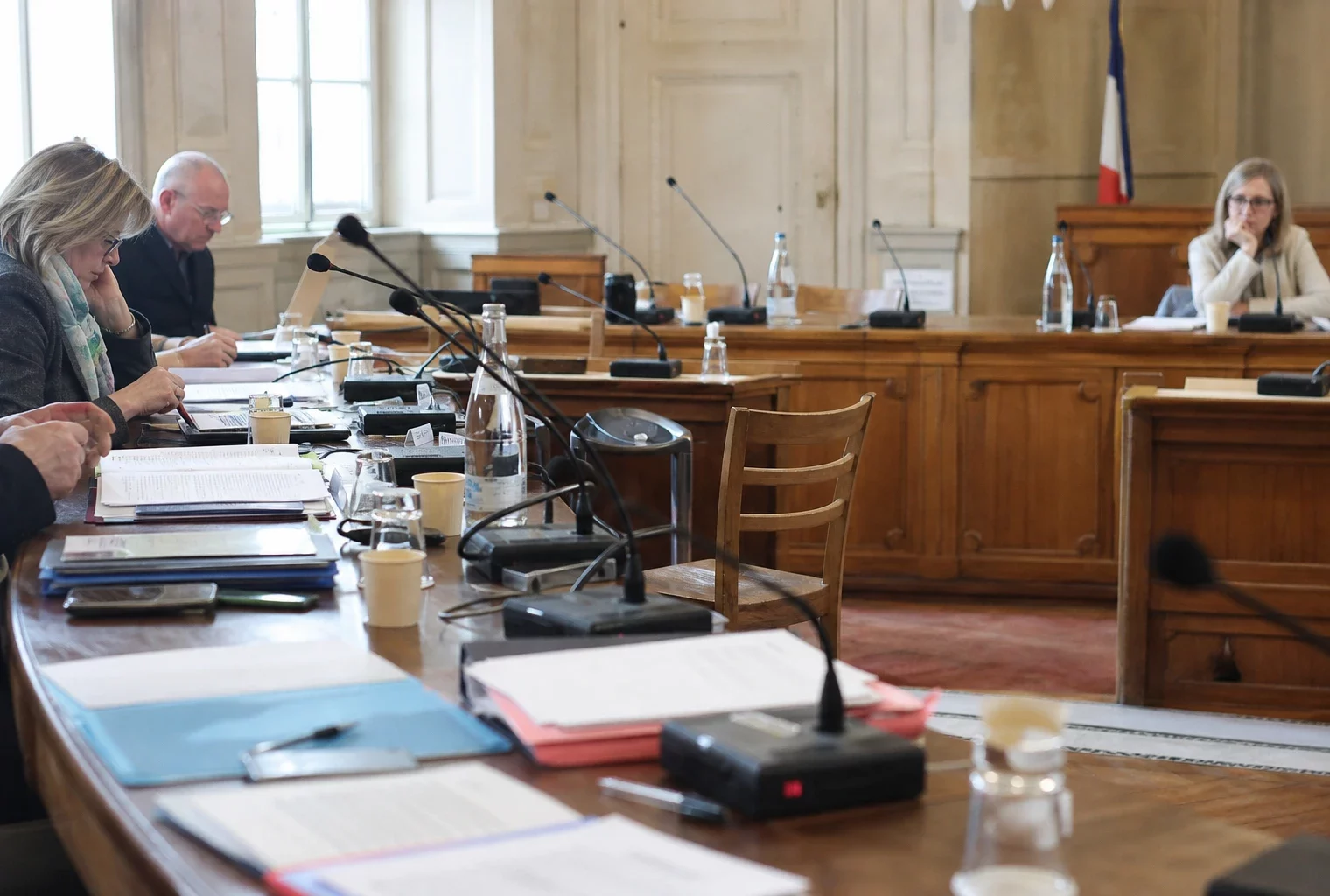 Salle du conseil municipal en lumière naturelle, avec chaise vide, micros et dossiers flous sur une table.