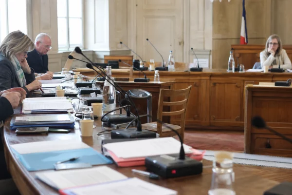 Salle du conseil municipal en lumière naturelle, avec chaise vide, micros et dossiers flous sur une table.