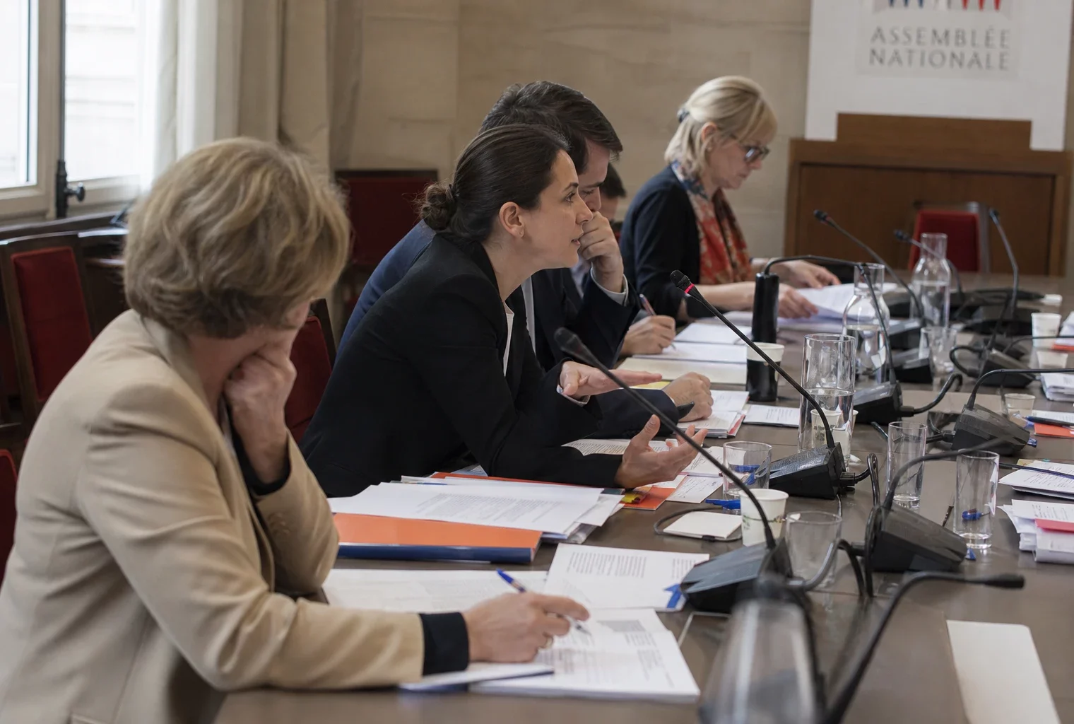 Réunion de commission à l’Assemblée nationale avec micros, dossiers ouverts et silhouettes anonymes autour d’une table.