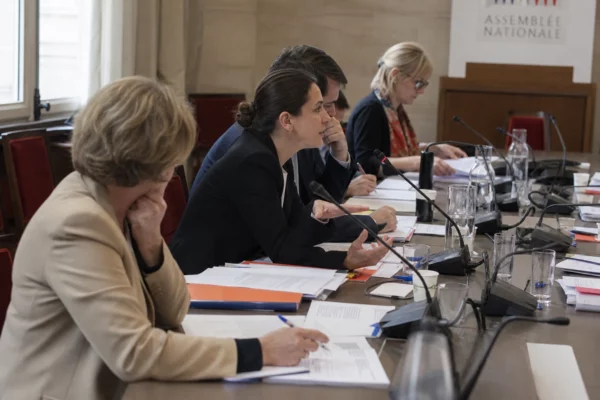 Réunion de commission à l’Assemblée nationale avec micros, dossiers ouverts et silhouettes anonymes autour d’une table.