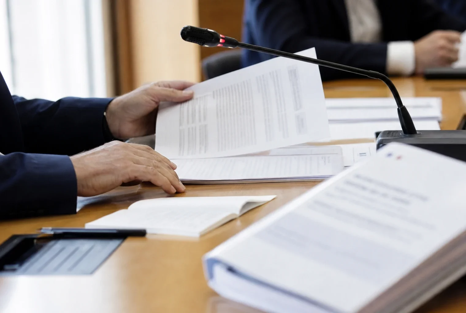 Mains anonymes consultant un dossier lors d’une audition institutionnelle, avec micro de commission et documents flous.