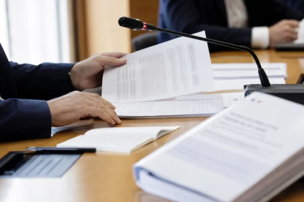 Mains anonymes consultant un dossier lors d’une audition institutionnelle, avec micro de commission et documents flous.
