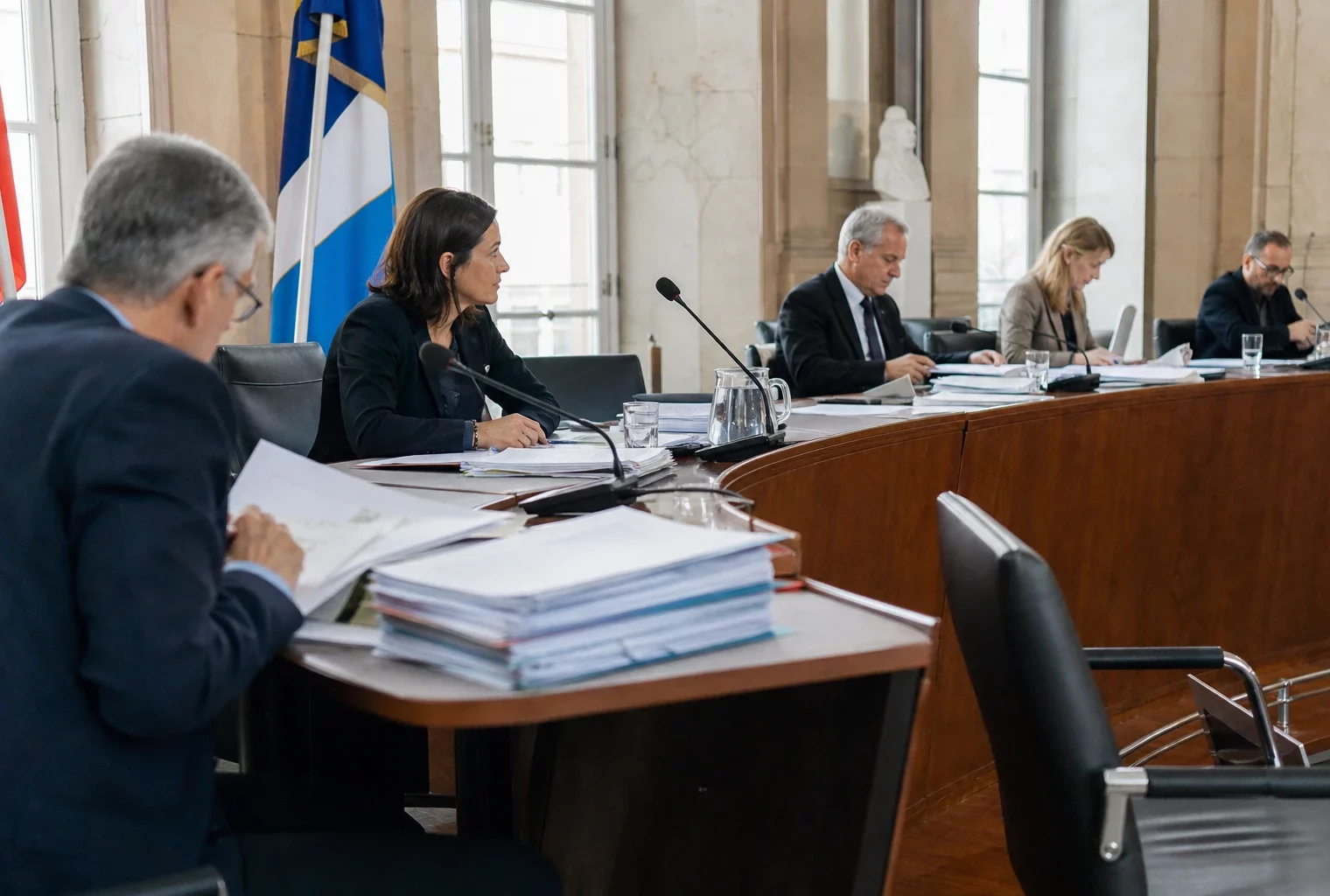 Réunion de commission dans une salle institutionnelle, micros de table, dossiers ouverts et élus anonymes sous une lumière claire.