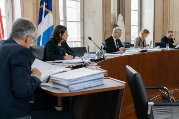 Réunion de commission dans une salle institutionnelle, micros de table, dossiers ouverts et élus anonymes sous une lumière claire.