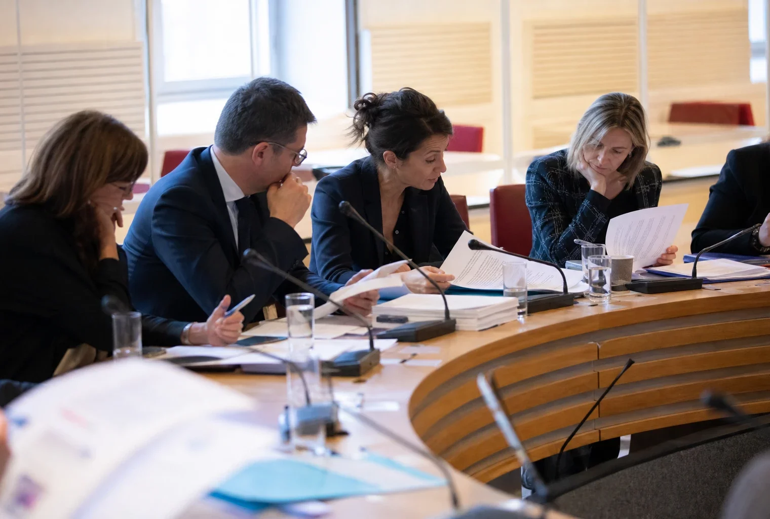 Réunion de commission parlementaire en France avec micros, dossiers ouverts et silhouettes anonymes sous une lumière claire.