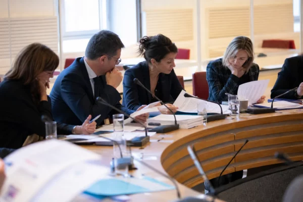 Réunion de commission parlementaire en France avec micros, dossiers ouverts et silhouettes anonymes sous une lumière claire.