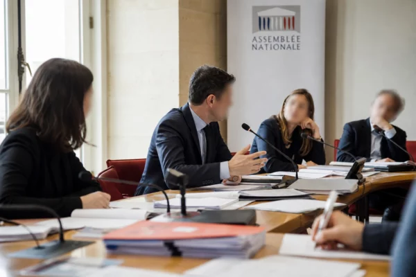 Réunion de commission parlementaire avec micros, dossiers ouverts et silhouettes anonymes sous lumière naturelle claire.