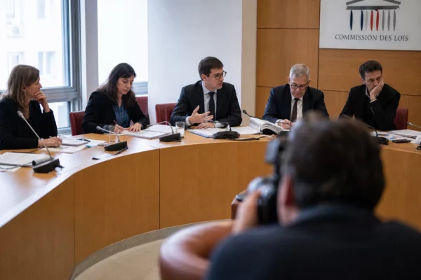 Réunion de commission parlementaire avec micros, dossiers ouverts et silhouettes anonymes dans une salle institutionnelle lumineuse.