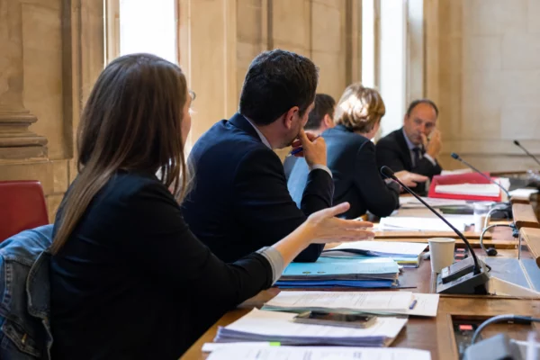 Réunion de commission parlementaire française avec micros, dossiers et silhouettes anonymes dans une salle lumineuse.