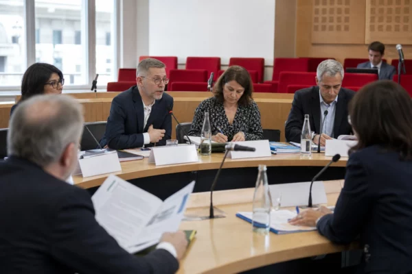 Réunion de commission parlementaire avec micros, dossiers et plusieurs participants anonymes dans une salle lumineuse.