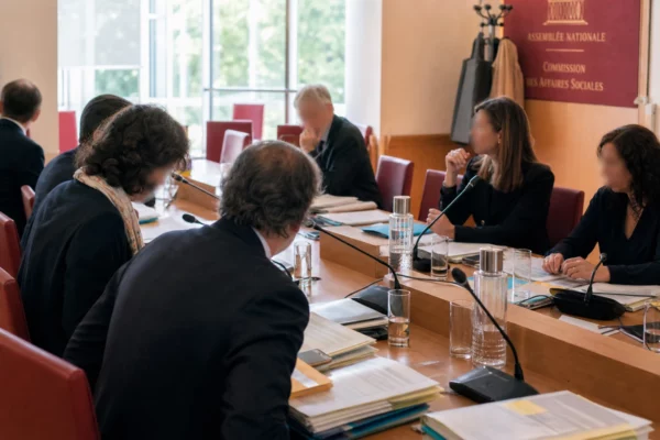 Réunion de commission parlementaire avec micros et dossiers, silhouettes anonymes dans une salle institutionnelle lumineuse