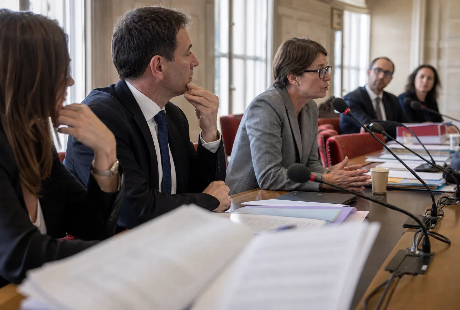 Réunion de commission parlementaire en France avec micros, dossiers ouverts et silhouettes anonymes sous une lumière claire.